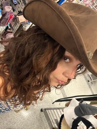 A woman with curly brown hair poses close to the camera, wearing a brown cowboy hat with a silver embellishment. She looks slightly upwards. In the background, there are retail shelves and a rack of hats including a white sun hat with a black ribbon, along with various packaged accessories. The floor is a light grey with speckles, suggesting a retail environment. The lighting appears to be bright and artificial.