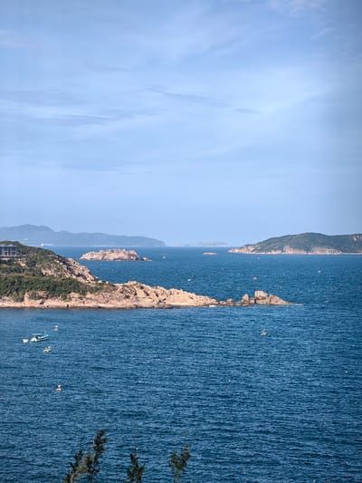 The scene depicts an elevated view of a coastal area in Tuy Phong 4, Vietnam, under a light blue sky with scattered cirrus clouds. The foreground features dark green foliage branches. The dominant element is a deep blue ocean, exhibiting ripples across its surface. On the left, a green, rocky headland juts into the water, supporting a multi-story building with a dark roof partially visible amidst vegetation. Two small vessels are positioned near this headland: a light blue fishing boat with a white cabin, and a smaller white boat adjacent to it. Numerous rocky islets and islands, varying in size and vegetation density, are distributed across the sea, extending toward the horizon. A larger, vegetated island is prominent in the right mid-ground. A solitary small white boat or kayak is situated in the open water to the right. Distant mountain ranges, softened by atmospheric haze, are discernible along the far horizon. Small white specks, consistent with buoys or remote objects, are scattered across the water surface.