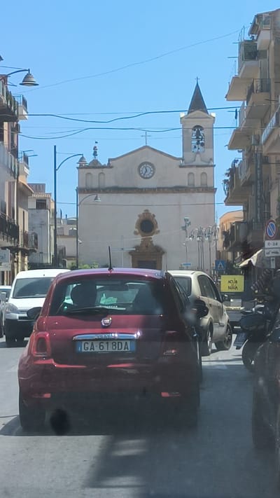 The image shows a street scene in Ficarazzi, Italy, with a church visible in the background. Several cars are on the road, including a red Fiat 500 in the foreground with the license plate "GA-618DA". The church has a clock tower with a cross at the top and bells visible. The surrounding buildings are multi-story structures with balconies. The sky is clear and blue, suggesting a sunny day. The scene appears to be a traffic situation, with vehicles stopped or moving slowly. There are also streetlights and power lines visible, indicating an urban environment.