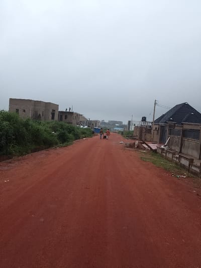 The image displays an unpaved red laterite road in Abeere, Nigeria, extending into the distance under an overcast grey sky. The scene is characterized by ongoing residential development and existing structures.
On the left side of the road, a dense strip of green vegetation borders the immediate roadside. Beyond this, a series of multi-story buildings are in various stages of construction. The closest prominent structure is a concrete block building with exposed rebar and multiple open window/door frames, lacking a finished roof. Three individuals are visible standing on the roof of this structure, indicating construction activity. Further down, additional uncompleted buildings are visible.
On the right side of the road, a completed or near-completed residential building is present, distinguished by its dark-tiled, pitched roof and a light-colored perimeter wall. A utility pole with overhead lines stands adjacent to this building. A black water storage tank is visible on a raised platform behind the property's perimeter wall. Scattered construction debris is visible on the ground near the roadside.
In the midground, on the red dirt road, two individuals are walking away from the camera. The individual on the left is wearing dark clothing. The individual on the right is wearing a lighter-colored top and darker trousers, and is pulling a light-colored wheeled cart or piece of luggage. The overall environment suggests a developing area with active construction.