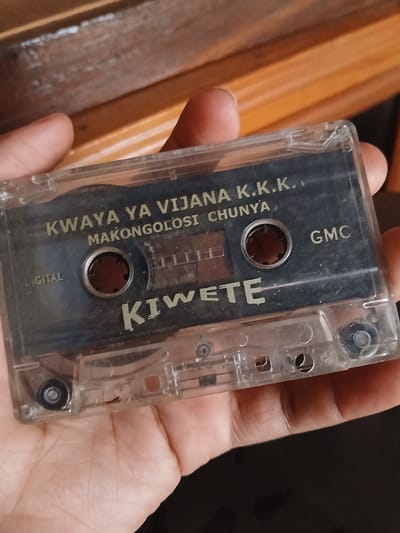 A hand holds a clear plastic audio cassette tape with a black label. The label features text in a gold-colored font. At the top, it reads "KWAYA YA VIJANA K.K.K." and below that, "MAKONGOLOSI CHUNYA". To the right of the text, "GMC" is visible. Below the central spindle hole, the word "KIWETE" is prominently displayed. To the left of the spindle hole, the word "DIGITAL" is partially visible. The cassette tape is held against a wooden surface, possibly furniture. The image does not contain information that would allow for determination of its location within Witeithie ward, Kenya.