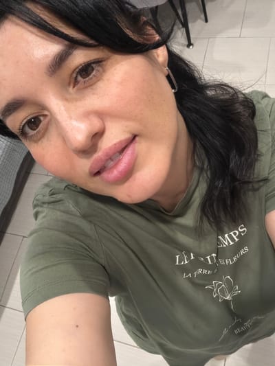 A close-up, overhead, indoor, selfie-style shot focuses on the face and upper torso of a woman with dark hair, dark eyes, and light skin with freckles. She is wearing a green t-shirt with white text and a floral design. The background is out of focus, but appears to be a tiled floor and possibly furniture. The woman is looking at the camera and smiling slightly. There is a rectangular silver earring visible in her left ear. There is no indication that this image was taken in Korolyov, Russia.