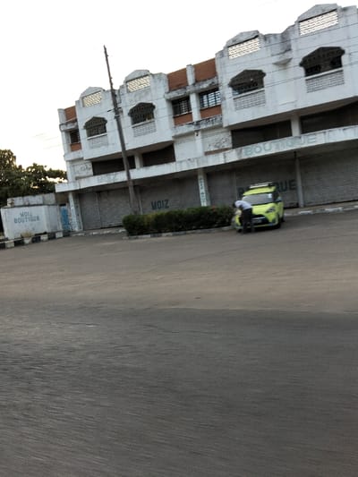 The image depicts an urban street scene in Gombato Bongwe ward, Kenya. In the background, a multi-story building with a light-colored facade, featuring reddish-brown sections and decorative elements such as patterned grates on its upper floor windows and roofline, is visible. The ground floor of this building consists of multiple commercial storefronts, all secured by closed metal roller shutters. Several shutters display "MOIZ" in dark lettering, while others show "BOUTIQUE." A utility pole stands centrally in front of the building. To the far left, a lower concrete structure also bears the inscription "MOIZ BOUTIQUE." In the mid-ground, a lime-green compact car, equipped with a roof rack, is parked facing the building. One individual, an adult male wearing a white shirt and dark trousers, is positioned in front of the vehicle, bent over towards its left front section. A low, trimmed hedge runs along the edge of the paved road, separating it from the building. The foreground comprises a wide asphalt road, marked by a black and white striped curb near the hedge. The sky above is clear and bright.