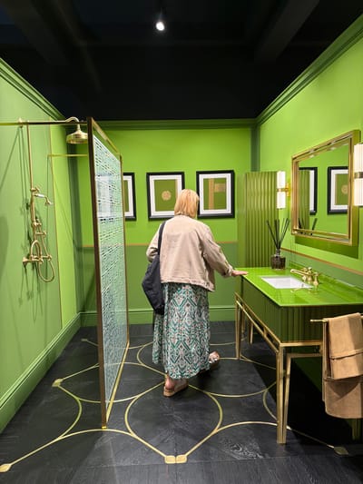 A woman is standing in a bathroom featuring green walls and a black floor with a gold geometric pattern. The woman has blonde hair and wears a light beige jacket over a long dress with a green and white pattern. She is facing a green vanity counter with a gold frame. The bathroom also includes gold fixtures, framed art on the wall, and a black doorway. This location is in Rho, Italy.