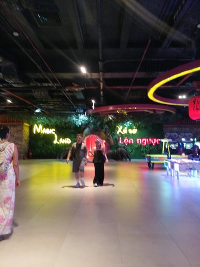 The image shows an interior scene in Hong Gai Ward, Vietnam, likely a shopping mall or entertainment center. In the foreground, a woman in a floral dress is partially visible on the left. Two people, a man and a woman, are walking in the center of the frame. The man is wearing shorts and a t-shirt, while the woman is dressed in dark clothing.
Behind them, a large sign reads "Magic Land" in yellow neon lights, situated against a backdrop of artificial greenery. To the right, another sign in Vietnamese is illuminated in red and green neon. Further back, there are arcade-style games and tables illuminated with bright neon lights. Overhead, the ceiling is exposed, revealing pipes, wires, and lighting fixtures. A circular neon light fixture hangs in the upper right. The floor is a light-colored tile, reflecting the various colors from the surrounding lights.