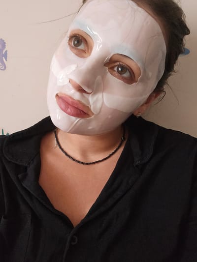 A close-up, selfie-style image shows a person wearing a white sheet mask on their face, with only their eyes and mouth visible. The mask is glossy and slightly translucent, with cutouts for the eyes and mouth. The person has brown eyes and full lips, visible through the mask's openings. They are wearing a black collared shirt and a thin, black beaded necklace with a small silver charm. The background is a plain, light-colored wall with a faint blue seahorse decoration on the left side. The lighting is soft and diffused, casting a slight shadow on the right side of the face. The image appears to have been taken indoors, possibly in a residential setting, and does not depict any specific landmarks or street scenes of Petropavlivska Borshchahivka, Ukraine.
