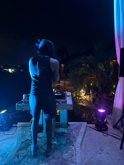 The scene depicts an outdoor nighttime event in Uluwatu, Indonesia, centered on an elevated stage where a DJ is performing.
A male individual, facing away from the viewer, stands centrally on a tiled platform, operating a DJ setup. He has long, dark hair exhibiting motion blur, wears a dark sleeveless tank top with a light vertical design on the back, dark shorts, and light-colored sneakers. His arms are raised, and his head is slightly tilted, indicating active engagement with the equipment.
The DJ setup consists of a rectangular, light-colored stone console. On the console are a DJ mixer and two media players, identifiable by their jog wheels and displays. A beverage can is visible on the left side of the console. The platform is illuminated by two stage lights: one on the left casts a prominent blue light onto the DJ and the stage, while another on the right emits purple light. A large dark speaker is partially visible on the far right, next to a flowing sheer white curtain illuminated by the purple light.
Below the elevated stage, a lush tropical environment is visible, featuring palm trees and other foliage illuminated by warm yellow ground lights. A body of water, possibly a pool or pond, reflects some of the ambient light directly in front of the stage. Further back, a group of indistinct individuals is gathered on a lower level, suggesting an audience or attendees at a social event. Buildings with illuminated interiors or exteriors are visible in the mid-ground and distant background under a dark night sky.