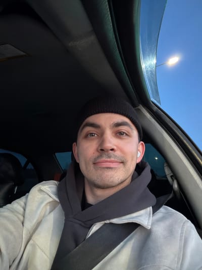 A selfie taken from inside a car. A man with a black beanie, wearing a gray hoodie and a white jacket, is looking at the camera. He has an earbud in his right ear. The car's sunroof is open, revealing a blue sky and a street light. The man is wearing a seatbelt. The location in Kuregovo, Russia, is not discernible from the image.