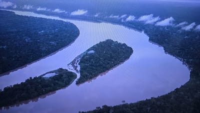 This aerial media file depicts a wide, meandering river flowing through a dense, dark green forest. The water appears muddy or silty, reflecting the overcast sky. There are several islands within the river, one larger than the others, covered in the same dense vegetation. Patches of lighter clouds are visible in the sky, suggesting a humid or rainy atmosphere. The overall scene is one of natural wilderness, likely a tropical or subtropical region given the dense forest cover. The time of day appears to be either early morning or late afternoon, as the lighting is diffused and shadows are not harsh. There are no people, objects, or visible text. The scene evokes a sense of remoteness and untouched nature. Based on the provided location context, this imagery does not align with Vlaardingen, Netherlands, as the landscape is characteristic of a vast river basin with extensive rainforests, not the typical Dutch environment.