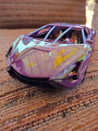 A close-up, low-angle, slightly blurry shot captures a small, iridescent purple toy car parked on a textured, brown woven surface, possibly a rug or upholstered furniture. The car's metallic finish reflects light in streaks of yellow, green, and blue. The car appears to be a sports car model, with a low profile and visible interior roll cage structure. The background is out of focus, showing muted colors that suggest an indoor environment with some natural light filtering in from the right. There are no people or animals visible in the image. The specific location in Galabovo, Bulgaria, is not discernible from the visual information provided in the image.