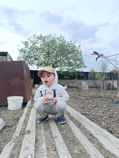 A child squats in an outdoor environment. The child wears a white hooded sweatshirt, gray sweatpants, and a tan baseball cap. The child's mouth is open in an expression of surprise. They are positioned on several parallel concrete blocks or beams. Behind the child is a garden or plot of land, a large tree with white flowers, and what appears to be a chicken coop structure with several chickens inside. To the left is a rusted metal container next to a white bucket. In the background, there is agricultural equipment. The photo was taken in Зыбинское сельское поселение, Ukraine.