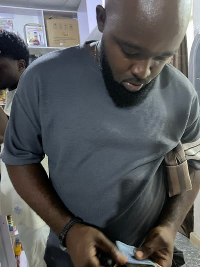 A man is seen in the foreground wearing a gray shirt with a brown and beige striped strap across his shoulder. He appears to be counting or handling what looks like money. He is dark-skinned, with a beard and a bracelet on his wrist.
In the background, there is a shelf filled with cardboard boxes, and other smaller items, suggesting a possible storage or retail environment. It is not possible to identify the specific location in Dushepe, Nigeria from the details provided in the image.