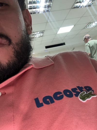 The image depicts an indoor scene in Adrogué, Argentina. The foreground shows the left profile of a person with a dark beard and mustache, visible from the upper lip area to the ear. This individual is wearing a pink polo shirt with a visible white button on the placket. The word "Lacoste" in dark blue text and its green crocodile logo are embroidered on the right chest area of the shirt. In the background, an older, bald male wearing glasses and a light green polo shirt is partially visible from behind, facing away from the camera and looking downwards. The ceiling features a white grid system with several recessed fluorescent light panels, some square with reflective louvers, and at least one rectangular flat panel, along with dark rectangular ventilation elements. A plain, light-colored wall is visible in the upper background.