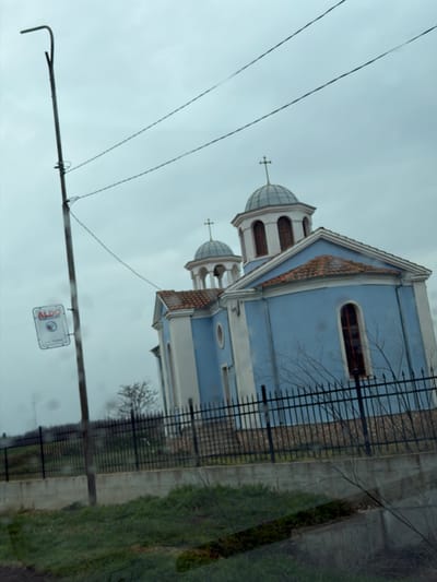 The image presents an exterior view of an Orthodox church located in Sveti Nikola, Bulgaria, under an overcast, light grey sky. The church building features pale blue walls with distinct white trim, brown terracotta-tiled roofs, and tall, dark-framed arched windows. Two light grey-domed structures are visible, each topped with a cross; a larger dome is positioned more centrally, with a smaller one behind and to its left.
In the left foreground, a dark utility pole with a curved top extends vertically, supporting multiple thin power lines that traverse the upper portion of the frame. Attached to this pole is a rectangular white sign displaying "ALDO" in red lettering along with a circular blue and white graphic below. A black metal picket fence runs across the midground, separating a patch of green grass and sparse, leafless branches in the foreground from the church property. Behind the fence, a concrete retaining wall is visible. In the far left background, a wind turbine is partially visible. No individuals or specific actions are observed in the scene. The overall environment suggests an overcast day in late winter or early spring.