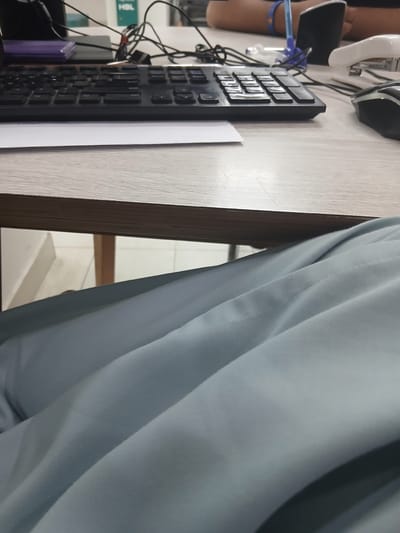 An indoor, eye-level, medium shot captures a desk with a keyboard, mouse, and stapler. The background is blurred, showing a person's arms and a blue pen. The foreground is dominated by a close-up of light blue fabric, possibly clothing, with visible folds and seams. The scene appears to be set in an office environment in Peshawar City Tehsil, Pakistan.