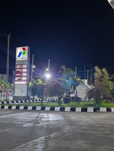 This is a night time, eye-level, exterior shot of a Pertamina gas station in Keuniree, Indonesia. The prominent feature is a tall, illuminated sign displaying the Pertamina logo and various fuel prices. The sign is lit by internal lighting, making the numbers and brand name clearly visible against the dark night sky. To the left of the sign is a utility pole with wires. In front of the sign, a paved area with a black and white striped border is visible, with grass and some potted plants. Behind the sign, palm trees and other foliage are illuminated by streetlights, creating a tropical atmosphere. A large white storage tank is visible in the background, along with more vegetation. The ground in the foreground appears wet, suggesting recent rain. There are no people visible in the scene.