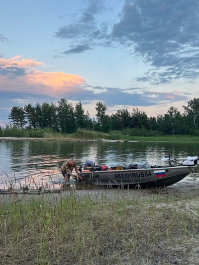 The image depicts a man kneeling by the water's edge next to a boat. The boat, likely a fishing or recreational vessel, is partially on land and partially in the water, indicating it might have just been docked or is about to be launched. The man is wearing camouflage clothing and appears to be engaged in some activity related to the boat or the water, possibly preparing gear or securing the vessel.

The scene is set by a calm body of water, with a sandy shore in the foreground. Beyond the shore, a dense line of trees forms the background, suggesting a natural, possibly rural or wilderness setting. The sky above is filled with dramatic clouds, with patches of blue sky and areas illuminated by the soft light of either sunrise or sunset, creating a serene and picturesque atmosphere. The light suggests it is either early morning or late evening.

Notable details include the boat's registration number "AB2301RUS54" printed on its side, along with a Russian flag decal. The man's attire suggests he is outdoors and possibly hunting or fishing. The overall mood is peaceful and quiet, capturing a moment of solitude in nature. The location context provided is Borovoye, Russia, which aligns with the natural scenery and the boat's registration.