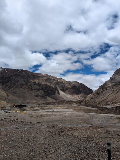 The image depicts a desolate, rocky landscape with towering, barren mountains under a sky filled with dramatic clouds. In the distance, a bridge spans a valley, and a few yellow tents are visible on the plain. A black lamp post stands in the foreground. The scene appears to be located in a high-altitude, arid region.