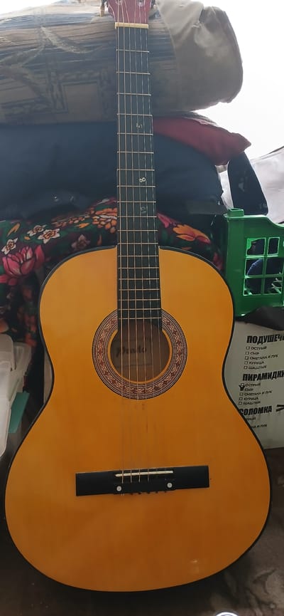 The image shows a yellow acoustic guitar standing upright, leaning against a stack of fabric items that appear to be bags or bedding. The guitar has six strings, a dark fretboard with white fret markers, and a circular soundhole with decorative trim. To the right of the guitar, a green plastic crate with a grid pattern is visible, and behind the guitar, there's a box with Russian text, possibly product packaging. The background consists of draped fabric and an off-white surface, suggesting an indoor environment, possibly a storage area or a room with clutter. The lighting is bright, casting subtle shadows. There are no people visible in the image. The location is specified as Mailuu-Suu City, Kyrgyzstan, but the visual elements themselves do not provide direct confirmation of this specific location, relying solely on the provided metadata.