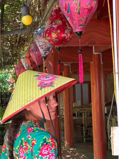 The image shows a vibrant scene in Thông Nguyên Commune, Vietnam, featuring a person looking upwards, surrounded by colorful lanterns. The individual, a woman, is seen in profile from the shoulder up, her face partially shaded by a traditional Vietnamese conical hat (nón lá). She has dark hair styled in braids, adorned with small white bead-like decorations. Her attire includes a striking green garment patterned with large red, pink, and yellow flowers.

The prominent nón lá she wears is yellow with a textured diamond pattern and a bold pink floral design. Above and around her, numerous lanterns of various colors and patterns hang from overhead structures. Notable lanterns include a pink one decorated with bamboo shoots and figures, and a red one featuring intricate floral motifs. Other lanterns, in shades of white, grey, and orange, also display diverse patterns and are often adorned with tassels.

The background consists of traditional wooden architectural elements, such as beams and posts, which provide a warm, reddish-brown contrast to the colorful foreground. Through gaps in the structures and surrounding foliage, hints of green trees and bushes are visible, suggesting an outdoor or semi-open setting. A single yellow light bulb hangs on a string near the lanterns. The scene appears to be set during the day, with natural light illuminating the area, enhancing the festive and decorative atmosphere created by the illuminated lanterns. No discernible text is visible within the image.