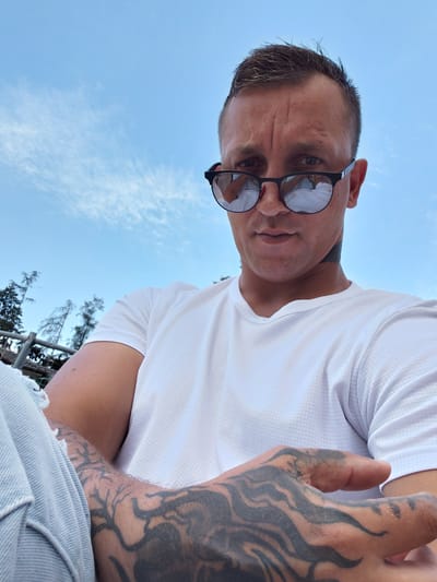 A man with short brown hair and a tattoo on his arm and hand is pictured outdoors. He is wearing sunglasses, a white t-shirt, and light-colored pants. The man is looking towards the camera with a serious expression. Behind him, the sky is blue with scattered clouds, and there are trees and a fence visible. The location is Levico Terme, Italy.