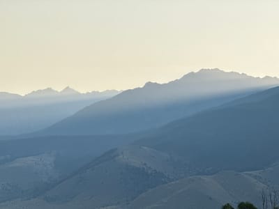 The image displays a panoramic landscape of mountainous and hilly terrain, observed from or near Livingston, United States. The upper third of the frame consists of a clear, pale yellow-beige sky. Below this, multiple layers of mountains and hills are arranged in receding planes, demonstrating atmospheric perspective. The nearest visible hills in the lower foreground exhibit undulating, sparsely vegetated slopes in lighter shades of blue-grey. Behind these, successive layers of larger mountains transition from darker blue-grey tones, suggesting denser tree cover, to lighter, hazier tones in the distance. A distinct diagonal band of light extends across a section of the mid-ground mountain range. The most distant mountains appear as faint, jagged silhouettes against the horizon, rendered in light blue-grey due to atmospheric haze. No human figures, man-made structures, or objects of human origin are present. The scene is characterized by soft, diffused illumination without a directly visible light source.