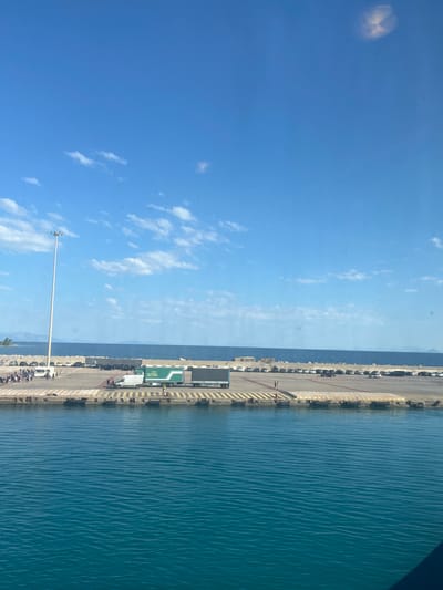 The image depicts a sunny day at the port of Kyllini, Greece. The foreground is dominated by the rippling, turquoise waters of the Ionian Sea. In the mid-ground, a concrete pier extends into the water, lined with yellow and black striped bollards. A tall, white lamppost stands prominently on the left side of the pier. Beyond the pier, a parking area is visible, filled with numerous cars and several commercial trucks, including a green and white truck with the logo "GVS". A group of people are gathered near the edge of the pier on the left, and a few individuals are scattered throughout the parking area. The background shows a clear blue sky with scattered white clouds, and a distant coastline with hills under the horizon. The overall scene suggests a busy port environment.