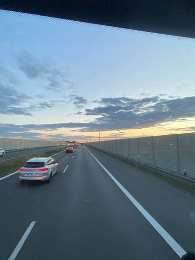 The image depicts a view from inside a vehicle on a highway near Węgrzce Wielkie, Poland, during twilight. The sky is a gradient of blue with scattered clouds, transitioning to a pale orange and yellow hue near the horizon. A sound barrier made of concrete panels lines the right side of the highway, stretching into the distance. On the highway, several vehicles are visible, including a silver station wagon in the foreground, its brake lights illuminated. The road is paved asphalt with clear white lane markings. Electric poles and wires are also visible along the side of the road. The overall scene suggests a normal traffic flow on a highway during the evening.