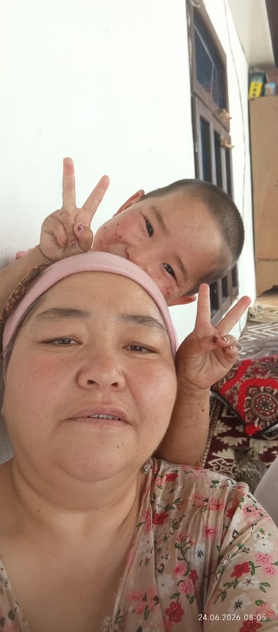 The image shows a selfie of two people, an adult and a child, likely mother and son. The adult, in the foreground, wears a pink headband and a floral-patterned shirt. The child, positioned behind and above the adult, has short hair and is making a peace sign with his left hand. He has some reddish marks on his cheeks. The adult also has her right hand raised, also making a peace sign. They are both looking at the camera. The background is a plain white wall, with a wooden door and window frame visible to the right. A patterned rug is visible on the right side of the frame, and a small cat can be seen sleeping on it. A timestamp in the bottom right corner indicates the date and time as 24.06.2026 08:05. The lighting suggests it is daytime, likely morning. The overall mood appears to be happy and playful, with both subjects smiling or looking engaged. The setting appears to be domestic, possibly indoors near a window.