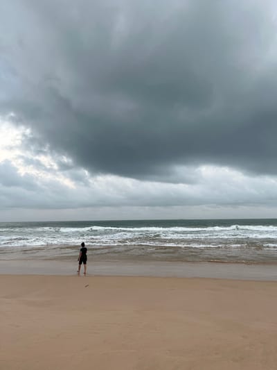 A lone individual stands at the edge of the water on a beach in Habaraduwa, Sri Lanka, facing the ocean under a dramatic, overcast sky. The person, seen from behind, is wearing dark shorts and a dark t-shirt with a blue collar. The sea is choppy with white-capped waves breaking onto the sandy shore. The sky is filled with heavy, dark gray clouds, suggesting an impending storm or recent rainfall. The sand is a light brown, appearing wet near the water's edge. The overall atmosphere is subdued and somewhat somber due to the weather conditions.