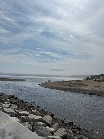 A wide-angle shot captures a serene coastal scene, likely in Magadan, Russia, under a hazy sky. In the foreground, a calm river flows from the right into a wider body of water on the left, its surface shimmering with reflected light. The riverbanks are lined with rough, grey rocks, some large and rounded, others smaller and more angular.
A narrow strip of sandy, pebble-strewn beach extends along the right side of the river. A few small groups of people, appearing as distant figures, are scattered along this beach, some walking, others standing. Beyond the beach, the hazy atmosphere obscures the distant landscape, revealing rolling hills or mountains veiled in a soft, white mist. A few structures, possibly buildings and tents, are visible on the far right of the beach, along with several parked vehicles.
The sky above is a pale blue, streaked with wispy, cirrus clouds that form delicate patterns across the expanse. The overall impression is one of quiet contemplation and the natural beauty of a coastal environment.