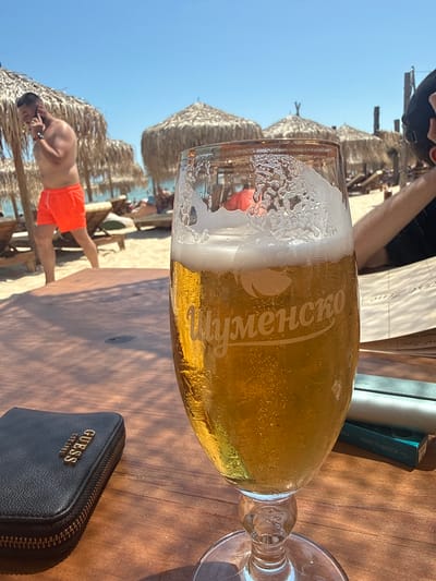 A man in orange shorts and a black t-shirt is walking on a beach in Kranevo, Bulgaria, while talking on his phone. In the foreground, a glass of beer sits on a wooden table, with a black wallet and some books in the background. The scene appears to be a beach bar or restaurant.