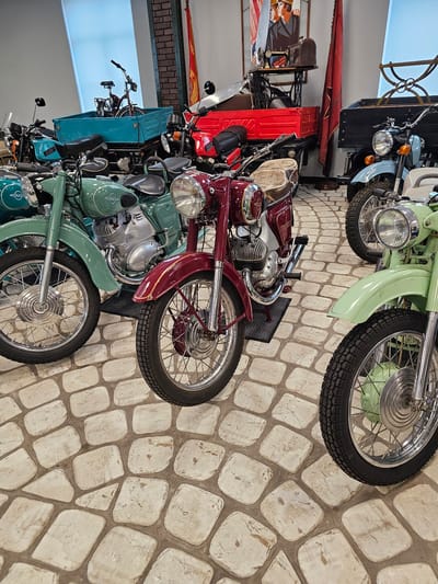 The image depicts a collection of vintage motorcycles displayed inside a building, likely a museum or showroom, in Архангельское, Russia. The motorcycles are various models, primarily in shades of light green and burgundy. The bikes are lined up closely together. In the background, other objects are visible, including what appears to be a vintage sewing machine, a red banner with a star, and other machinery. The floor is paved with light-colored stone.