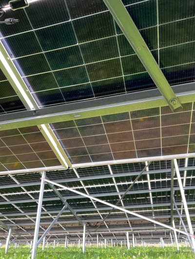 The image shows the underside of a solar panel array. The structure is constructed of galvanized steel beams and supports. The panels themselves are rectangular, and their backs are visible, displaying a grid-like pattern of individual solar cells. The setting is outdoors, with green grass visible at the edge of the frame, suggesting the location is likely a solar farm or an open area on the outskirts of Grange Ville, Ireland. There are no people visible in the image, and the focus is entirely on the technical structure of the solar panels.