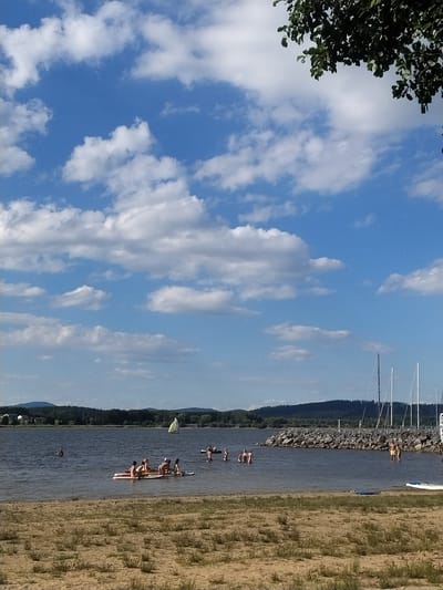 A bright, sunny day is depicted at a lake in Radslav, Czechia, with a clear blue sky dotted with fluffy white clouds. In the foreground, a sandy beach is sparsely covered with dry grass. The middle ground features the calm, dark water of the lake, where several groups of people are enjoying themselves. One group of about six individuals is lounging on a light-colored inflatable raft, appearing relaxed. Another group of approximately four people are standing in the shallow water near the raft. Further out in the lake, a lone person is swimming, and in the distance, a small yellow sailboat is visible. Along the far shore of the lake, a dense line of dark green trees forms a natural barrier, with a few scattered houses peeking through. To the right side of the image, a stone breakwater juts out into the water, and several white sailboat masts are visible docked behind it. The overall scene conveys a peaceful and recreational atmosphere on a warm summer day.