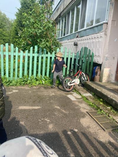 This image depicts an outdoor scene in Ihnatpil, Ukraine, featuring a young boy, a bicycle, and a residential building under natural light, casting prominent shadows.
In the mid-ground, a young boy, approximately 6-8 years old, is visible. He wears a camouflage bucket hat, a dark t-shirt, gray sweatpants, and dark sandals. He is positioned slightly left of center, with his left leg raised and right foot on the ground, indicating movement. He looks towards the right side of the frame.
To the boy's right, a red, black, and white children's bicycle, branded "ARDIS" on its frame, is parked on a concrete path, leaning against a light green wooden picket fence. The bicycle has red rims. The fence extends across the middle of the frame, bordering an overgrown garden area with various green plants and some orange flowers.
Behind the fence, a lush green bush with dark leaves is prominent, with taller trees and a brighter sky in the background. To the right of the fence, a multi-story residential building is visible. Its facade is a faded pinkish-beige with a textured pattern of circular indentations on the lower wall. Above this, a long, enclosed balcony or series of windows with white frames extends across the building. Below the windows, a metal vent or pipe fixture protrudes from the wall. Near the ground level, to the right of the bicycle, a small, dark rectangular opening is present in the building's foundation, with a wooden log or stump and a rusty metal barrel nearby. A blue cloth hangs near this area.
The foreground consists of a gray, asphalt-like concrete ground surface. Long, striped shadows from the fence pickets are cast onto the ground, indicating direct sunlight from the left or right. In the bottom left corner, part of a white and blue football with the word "SANTANA" partially visible is in extremely close foreground, suggesting its proximity to the camera or observer. Two metal drainage grates are present on the ground: a smaller one to the left and a larger, rectangular one to the right, adjacent to a raised concrete curb or ramp that runs along the right edge of the path, bordered by weeds and grass.