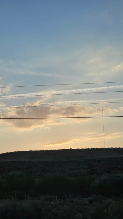 A photograph captures a vast expanse of sky at dusk, with horizontal power lines traversing the frame. Below the sky, a dark, undulating silhouette of a hill or ridge is visible, sparsely dotted with low-lying vegetation. The sky transitions from a clear, pale blue at the top to a warmer, golden hue near the horizon, where scattered clouds catch the fading sunlight. Streaks of contrails from aircraft are prominent across the upper and middle sections of the sky. A tall, thin tower, possibly a radio or cell tower, stands on the right side of the ridge. The foreground is dominated by dark, shadowed foliage and grass. The image does not depict any individuals or specific actions, but rather a natural landscape under a twilight sky. The provided context suggests this scene is located in the city of La Muela, Spain.