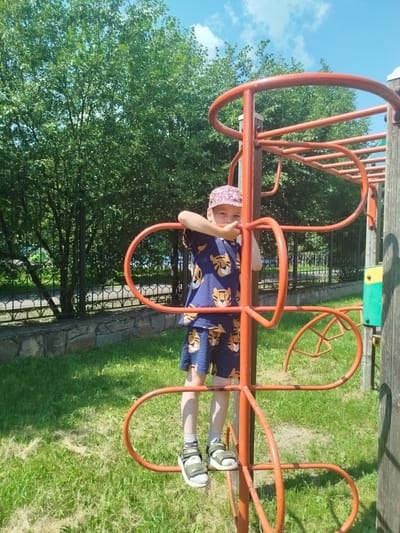 A young boy stands on a playground climbing structure in Yalutorovsk, Russia. He is wearing a blue t-shirt and shorts with a tiger pattern, a patterned cap, and sandals. The boy is holding onto the orange metal bars of the structure, which is set against a backdrop of green trees and a blue sky. The playground appears to be in a park or outdoor recreational area.