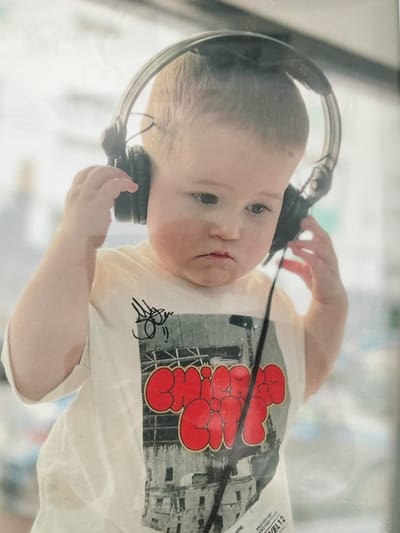 A young child is wearing headphones and a t-shirt with a graphic design. The child's hands are near the headphones, suggesting they are adjusting them or holding them in place. The t-shirt has a large red graffiti-style word on it, overlaying a black and white image of buildings. The background is blurred and out of focus, but appears to be a brightly lit interior or exterior space. There is a dark cord extending from the headphones down the child's shirt. The child's expression is slightly serious or contemplative. No specific city or location can be determined from the image itself.