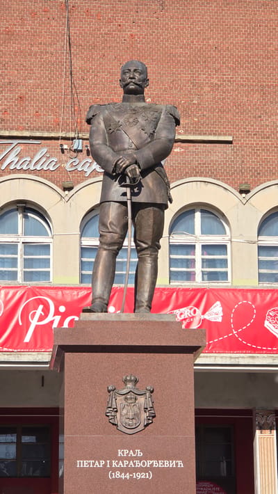 The image displays a bronze statue of King Peter I Karađorđević (1844-1921) in an urban setting, specifically in Subotica, Serbia. The king is depicted standing, wearing a military-style uniform adorned with numerous medals and epaulets, and holding a saber. He has a mustache and a serious expression.
The statue rests on a red-brown granite pedestal. On the front of the pedestal, there is a relief of the Royal Coat of Arms of Serbia/Yugoslavia, flanked by text.
Behind the statue is a multi-story brick building featuring multiple arched windows, some round and some rectangular with arched tops. A large red banner, possibly an advertisement or event announcement, covers a significant portion of the building's facade, partially obscuring the windows on the lower levels. Another sign, partially visible on the left, reads "Thalia cap" in cursive. In the background on the right, text visible through a window confirms the presence of a "НАРОДНО ПОЗОРИШТЕ" (National Theatre).
The scene is captured on a bright, sunny day, indicated by the clear shadows cast on the statue and the building. No specific activity or event is taking place, but rather the image captures a monument in its urban environment.