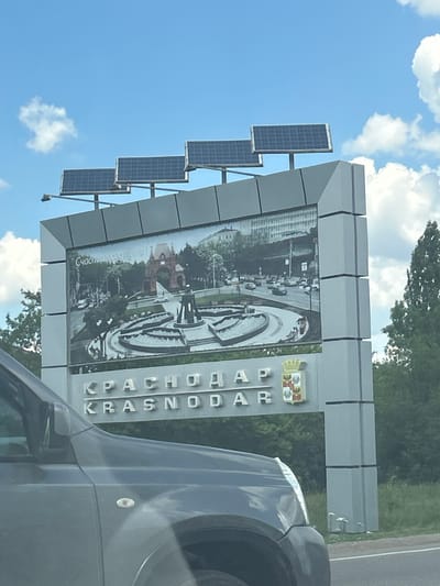 This media file depicts a large, modern sign for Krasnodar, Russia, positioned near a road. The sign features a large photograph of a city scene, possibly a fountain and surrounding buildings, set against a bright blue sky with scattered white clouds. Below the photograph, the word "КРАСНОДАР" is prominently displayed in large, silver, three-dimensional letters. There is also a coat of arms displayed on the right side of the sign.

Attached to the left side of the sign structure are several solar panels, suggesting an eco-friendly power source for the sign's illumination or other functions. The overall scene appears to be captured during the daytime, with clear weather conditions. The perspective of the shot suggests it was taken from a vehicle, indicated by the presence of a car's fender and a portion of its bodywork in the foreground, partially obscuring the sign. The image is rotated, with the sign and sky appearing vertically oriented. The setting appears to be an approach to or exit from the city of Krasnodar.