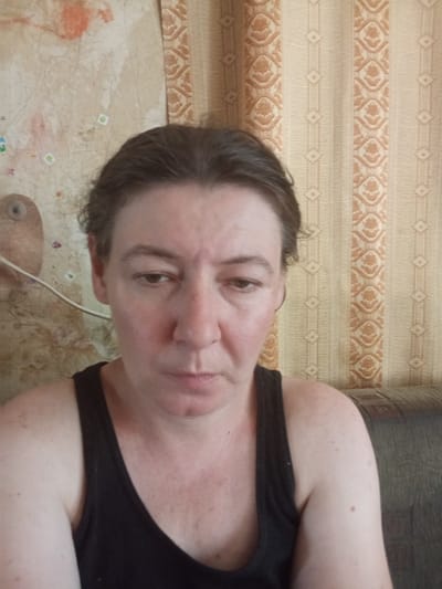 A mid-shot, selfie-style photograph shows a woman with dark hair pulled back from her face, looking downwards. She is wearing a black tank top and has fair skin with some freckles. Her expression is somber. The background consists of a wall with a textured, patterned wallpaper in beige and orange hues on the right side and a different, lighter patterned wall on the left. A piece of white cable is visible on the left side of the image. The setting appears to be an indoor residential environment in Berezivka, Ukraine.