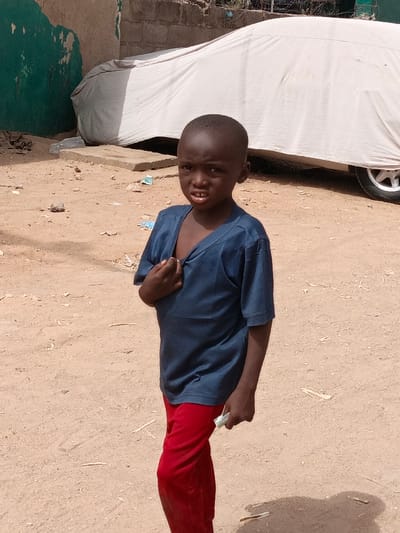 An image taken in Zango, Nigeria, depicts a young dark-skinned child standing outdoors on a light brown, sandy ground. The child, dressed in a dark blue short-sleeved shirt and red pants, is positioned centrally, looking directly towards the camera with a slight frown and visible teeth. The child's right hand grips the collar of the shirt, pulling it slightly open, while the left hand holds a small, light-colored object, possibly folded currency or paper. In the background, a light-colored vehicle, likely a car, is completely covered with a white or off-white tarp, with its rear passenger-side wheel (silver rim) partially visible on the right. To the left of the covered vehicle, a weathered wall structure with a faded green-painted section is visible. Scattered debris, including small pieces of plastic and other refuse, lies on the dusty ground. A concrete slab or block is situated behind the child, between the wall and the covered vehicle. The scene is brightly lit by natural sunlight, indicated by visible shadows.