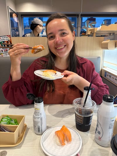 In the foreground of this medium shot, a Caucasian woman with long dark hair and fair skin smiles at the camera while holding a piece of sushi with chopsticks. She is wearing a dark red, long-sleeved shirt. In front of her on a light-colored table, there is a small white plate with two pieces of salmon sushi, a clear plastic cup with a dark beverage and a black straw, a bottle of soy sauce, and a small beige container filled with green packets. Behind her, a conveyor belt sushi restaurant can be seen through a window, with patrons and employees visible. The restaurant appears to be in the city of Chiyoda, Japan, based on the visible text on signs and packaging. The overall lighting suggests it is either dusk or indoors with artificial lighting.