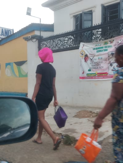 In Iju, Nigeria, a full shot captures a street scene with two women walking. The woman on the left, in the foreground, wears a pink headscarf and a black t-shirt with black shorts, carrying a purple bag in her right hand. She is walking away from the camera, and her left hand is out of view. The woman on the right, partially visible, wears a patterned dress and carries an orange bag in her right hand. She is walking towards the left of the frame, with her left arm also out of view. The background features a white building with dark-trimmed windows, a black decorative fence, and a banner that appears to be a campaign poster with an image of a man and text, including "VOTE!". The ground is a dirt and concrete surface with some debris. A portion of a car's side mirror is visible in the lower left of the frame, reflecting a cloudy sky and a road.