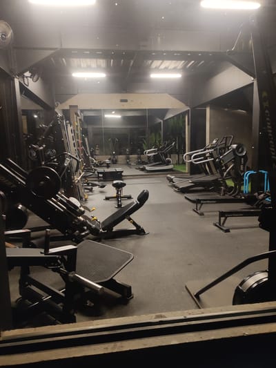 The image displays an unoccupied indoor gym facility, located in Mas, Indonesia. The perspective is from a low vantage point, possibly through a window or doorway frame. The gym features a dark grey, possibly rubberized floor. The walls are dark, with a significant portion on the right consisting of a large mirror that reflects the interior of the gym and an exterior view of green foliage seen through an adjacent glass partition. The ceiling is dark, revealing structural beams, and is equipped with two circular wall-mounted fans. The space is brightly illuminated by multiple linear overhead fluorescent or LED strip lights.
Various fitness machines populate the room. In the left foreground, a black Hoist leg press machine with circular weight plates is prominent, with part of a Nautilus machine visible below the frame. Moving towards the background on the left, several strength training machines and a black adjustable utility bench are arranged. On the right side, multiple cardio machines are present, including two black treadmills (one labeled "Hittube Tmill," another "Speedfit"), and at least two StairMaster brand rowing machines. A blue metal frame, likely part of a squat rack or storage, stands near the cardio equipment. A Concept2 rowing machine is partially visible on the far right. The mirror reflects additional gym equipment in the background, such as a squat rack with yellow resistance bands and an air bike, alongside a section of green artificial turf. No individuals are present, and no actions or interactions are depicted.