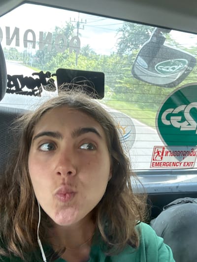 An image captured from inside a vehicle depicts a young adult female in the foreground, with the exterior environment visible through the rear windshield. The scene is located in Khlong Thom, Thailand, confirmed by the presence of Thai script on an emergency exit sign.

The female, positioned in what appears to be a back seat, occupies the lower half of the frame. She has brown, wavy hair, prominent eyebrows, and is wearing a green top and white wired earbuds. Her expression features pursed lips and her eyes are directed upward and to the right.

The rear windshield of the vehicle is adorned with several stickers and objects. Visible from the inside, these include reversed white text, partially reading "RONANO," on the upper left, and a black silhouette sticker below it. On the right side, a large black object with a reflective surface and a logo, a green circular sticker featuring a reversed "CGS" or "CES" logo, and a smaller blue and yellow wave-motif sticker are present. A red rectangular sign with white text stating "EMERGENCY EXIT" and its Thai equivalent, "ทางออกฉุกเฉิน," is affixed to the lower right of the windshield.

Through the rear windshield, a paved road is visible, curving to the right, flanked by dense tropical vegetation and trees. Utility poles carrying multiple power lines line the left side of the road. The sky is bright with scattered clouds. Inside the vehicle, a dark, lumpy object, consistent with a bag or backpack, is partially visible in the lower right foreground.