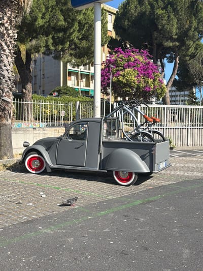 A gray, two-door Citroën 2CV pickup truck is parked on a paved surface in an urban setting. The truck has a modified cargo bed in the rear, carrying two bicycles. The front wheel has a red rim and white tire. The vehicle's license plate is partially visible, reading "EB 762FB". A pigeon stands on the cobblestone ground to the left of the truck. In the background, a white metal fence separates the parking area from a sidewalk or walkway. Beyond the fence, there are buildings and lush green trees, including a tree with vibrant pink blossoms. The overall scene suggests a sunny day. The image was taken in Vallecrosia al Mare, Italy.