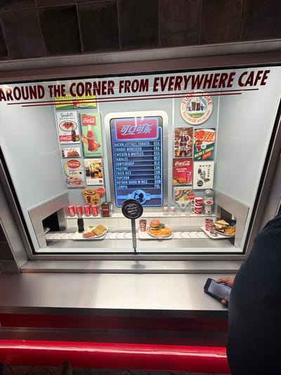 A vertically oriented, medium shot features a display case for the Coca-Cola Company cafe "Around The Corner From Everywhere Cafe," potentially located in Atlanta, USA. The exterior of the case is constructed of gray metal and glass. An individual is partially visible at the top right of the frame, wearing a dark blue shirt and holding a black smartphone.
Inside the display case, there is an assortment of Coca-Cola themed vintage advertisements and menu items. The menu board, located in the center, lists various food items and their country of origin. The advertisements showcase vintage Coca-Cola ads in various languages, alongside representations of food items and Coca-Cola products. Replicas of food items and Coca-Cola beverages are arranged on a miniature counter, mimicking a cafe serving station.
The background is nondescript. A dark wall is seen on the left, and a red structure appears on the right.