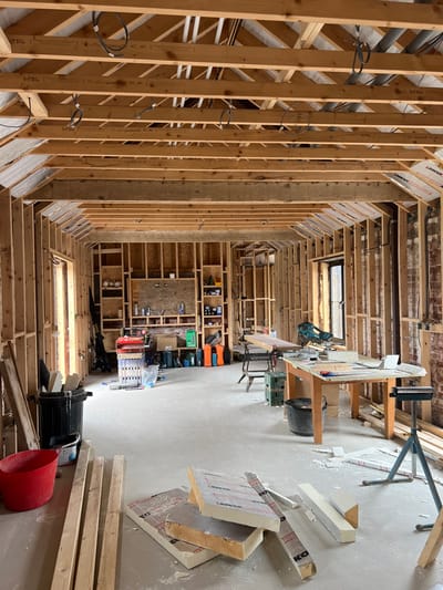 The image depicts an interior construction scene within a building located in Skipsea, United Kingdom. The space is currently undergoing significant renovation or build-out, characterized by exposed timber framing for walls and the ceiling/roof structure.
The environment features a light grey concrete or screed floor covered with construction debris, including sawdust and fragments of insulation. The walls are primarily framed with light-colored timber studs, with sections of older exposed red brickwork visible behind the framing on the right side. The ceiling shows a complex network of exposed timber joists and rafters, with some electrical wiring visible and sections of foil-faced rigid insulation panels attached to the underside of the sloped roof on the right.
No individuals are present in the scene.
Objects within the space include:
* In the foreground left, a red plastic bucket, a black refuse bin, and several cut timber planks are on the floor.
* In the foreground right, multiple pieces of rigid foam insulation panels are stacked on the floor, along with scattered insulation fragments and sawdust. A metal tripod stand is also present.
* In the midground, towards the left wall, a section of timber framing is configured as built-in shelving, holding various bottles, cans, and small items. Below this shelving, assorted power tools in cases and orange containers are on the floor. A red metal multi-drawer tool caddy is positioned nearby, alongside a black hand truck.
* A wooden work bench supported by black sawhorses is in the midground center-left, with a large piece of wood on its surface.
* A green plastic crate rests on the floor near the workbench.
* In the midground right, a large wooden work table is positioned, covered with sawdust and insulation fragments. A teal-colored miter saw is set up on this table, facing right. Various hand tools and materials are scattered across the table surface. A black bucket is situated beneath the table.
* Framed openings, likely for windows or doors, are visible on the left and right walls.
The presence of construction materials, tools, and debris indicates active or recently ceased construction or carpentry work within the building.